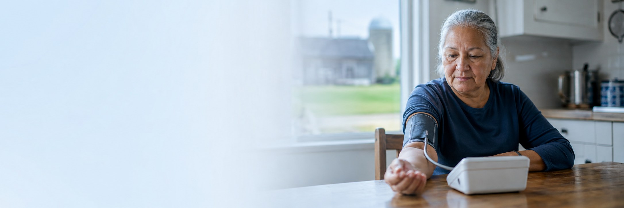 Older woman taking a home blood pressure reading at her kitchen table, a barn and silo visible through the window